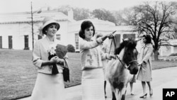 Në këtë fotografi të 12 prillit 1962, zonja e parë amerikane Jacqueline Kennedy i bën një turne udhërrëfyes në ambientet e Shtëpisë së Bardhë perandoreshës Farah Pahlavi të Iranit.