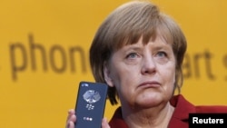 Angela Merkel arătându-și telefonul mobil (“Handy”) în octombrie 2013, când s-a dovedit că serviciile secrete din SUA au putut monitoriza telefoanul cancelarei germane. Despre denumirea germană de “Handy” pentru un telefon mobil — mai multe aici, în revista presei.