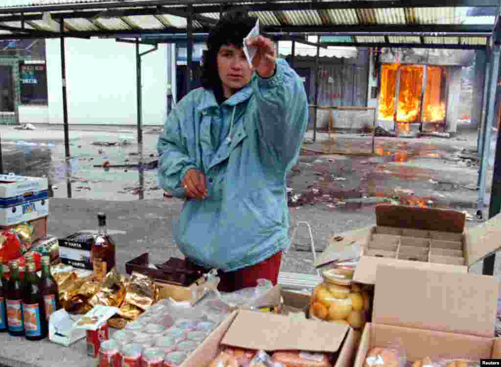 Serb aýal bazarda zat satýar, onuň arkasynda Ilidziada, Saraýewonyň serbleriň elindäki eteginde ýanýan dükan görünýär. 9-njy mart, 1996. 