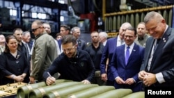 Ukrainian President Volodymyr Zelenskiy (signing a shell in the center of the picture) visits a munitions manufacturing plant with Ukrainian Ambassador to the United States Oksana Markarova in the background (far left).