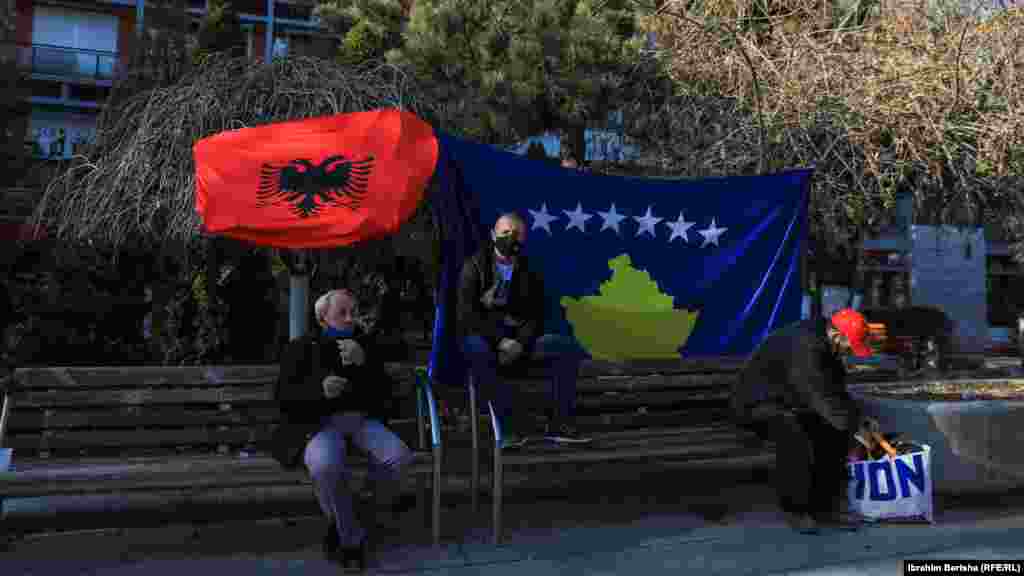 Disa qytetarë janë ulur në sheshin “Nëna Terezë”, prapa tyre qëndron flamuri i Kosovës dhe ai i Shqipërisë.